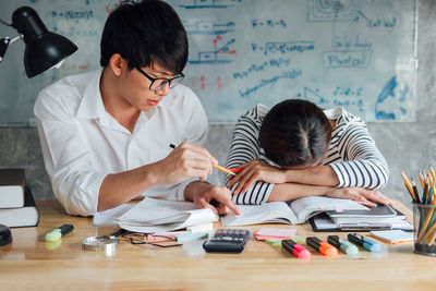 Young man reading book by exhausted friend napping on desk