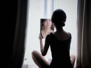 Rear view of woman looking at mirror sitting by window