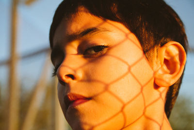 Close-up of boy standing with chain link fence shadow on face outdoors