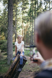 Smiling daughter sitting on log while father photographing through smart phone in forest