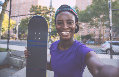 Portrait of smiling young woman holding skateboard standing in city