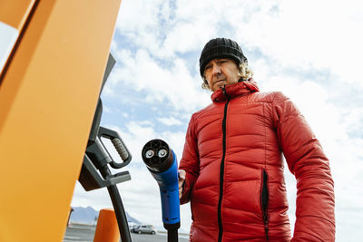 From below of thoughtful male in warm outwear and hat standing with charging power to electric vehicle in hand at gas station in sunny day