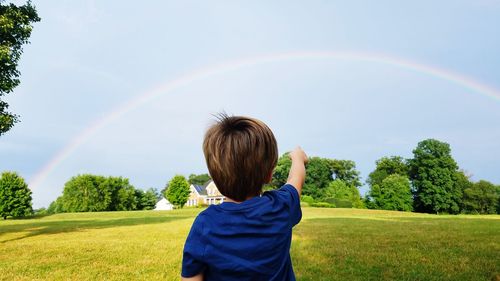 Rear view of boy pointing at rainbow while standing on field