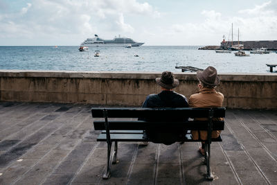 Rear view of people sitting on bench by sea against sky