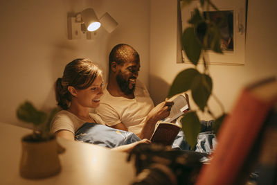 Smiling couple reading books by illuminated sconce mounted on wall at home