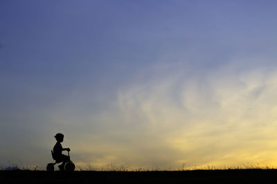 Silhouette girl riding bicycle against sky during sunset