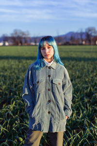 Thoughtful young female with blue hair looking at camera dressed in trendy jacket standing in green field in sunny evening
