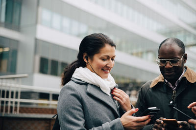 Smiling colleagues using mobile phones while standing against building in city