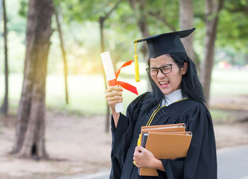 Young woman with eyes closed holding certificate and books while standing on road against trees