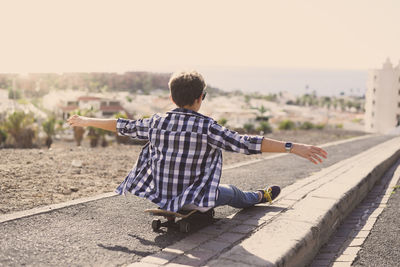 Rear view of boy with arms outstretched sitting on skateboard at footpath