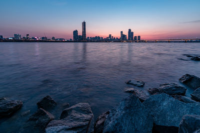 Sea by city buildings against sky during sunset