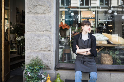 Young female owner with arms crossed sitting on store window sill