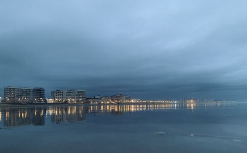 Illuminated buildings by sea against sky at night