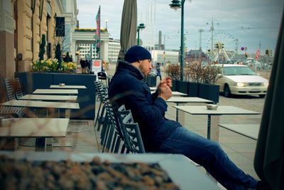 Man smoking cigarette while sitting on chair at sidewalk cafe in city