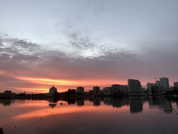 Silhouette buildings by river against sky during sunset