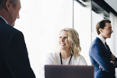 Happy female entrepreneur talking to colleague while coworker standing in background at workplace