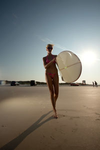 Mid adult woman with surfboard walking at beach against sky