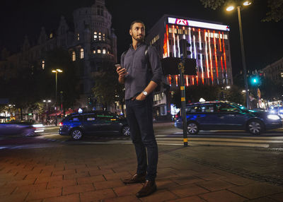 Full length of man looking away holding smart phone in city at night