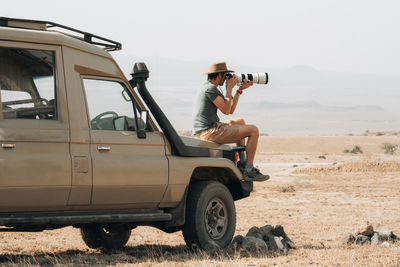 Side view of male traveling photographer sitting on offroader and taking picture on camera with telephoto lens during safari in savannah in summer