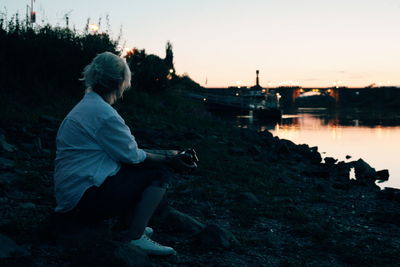 Man sitting at lakeshore against sky during sunset