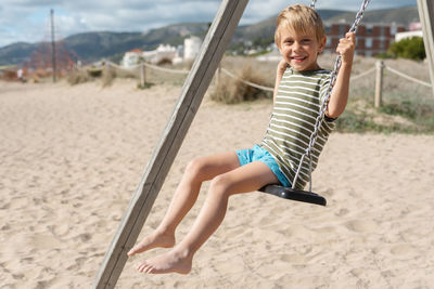 A happy boy on a swing on a sandy beach against the backdrop of apartment buildings and a stormy sky