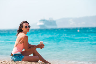 Young woman sitting on sunglasses at beach against sky