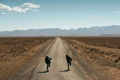 Rear view of friends skateboarding on road amidst field against sky