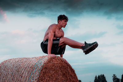 Low angle view of shirtless teenage boy exercising on hay bale against sky