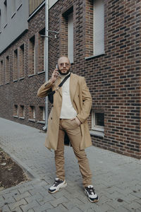 Portrait of man talking on phone while standing against building on footpath