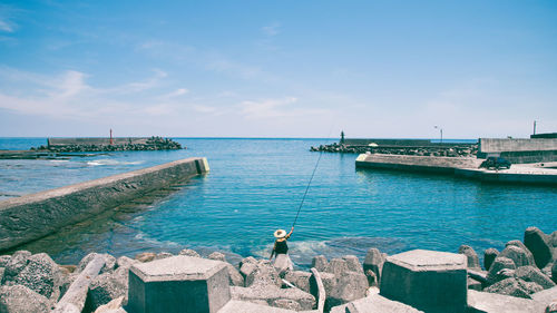 Rear view of person fishing in sea while sitting on groyne against sky