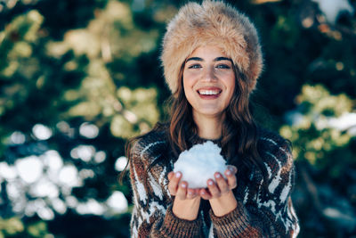 Portrait of young woman holding snow while standing against trees during winter
