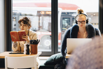 Customers reading novel and using laptop against window at coffee shop