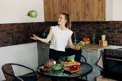 Cheerful young woman preparing green salad at home and jokingly throwing vegetables up