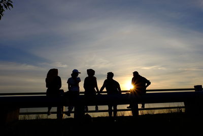 Silhouette friends sitting on railing against sky during sunset