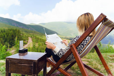 Woman sitting on chair by mountains against sky