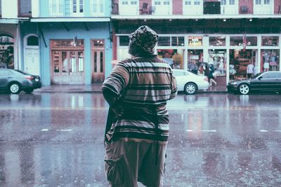 Rear view of man with hand on hip standing on footpath during rainfall