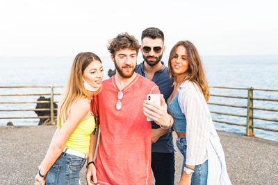 Cheerful friends doing selfie while standing against sea