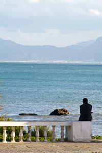 Rear view of man sitting on retaining wall while looking at sea against sky