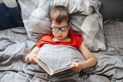 Boy with glasses lies on a bed under a blanket and reads a book, top view. 