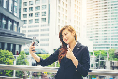 Smiling young businesswoman showing thumbs up while taking selfie against in city