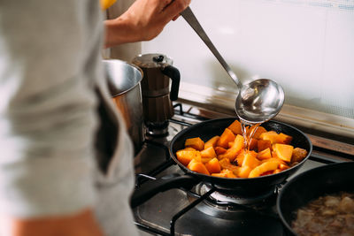 Crop anonymous woman with kitchen spoon pouring water into frying pan with chopped pumpkin while preparing cream soup