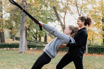 Happy friends exercising with straps while standing by tree