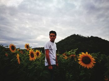 Young man standing on sunflower farm against cloudy sky