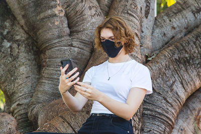 Midsection of man using mobile phone on tree trunk