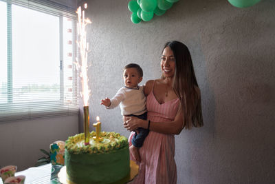 Delighted ethnic mom holding baby boy while celebrating birthday with festive cake decorated with burning firework