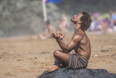 Shirtless young man gesturing while meditating on rock at beach during summer vacation