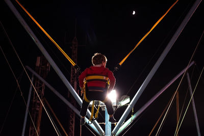 Low angle view of boy on trampoline against illuminated lighting equipment at night