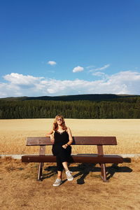 Full length of woman sitting on bench against blue sky