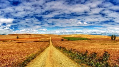 Dirt road passing through field against cloudy sky