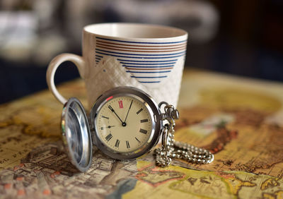 Close-up of pocket watch with cup on map at table